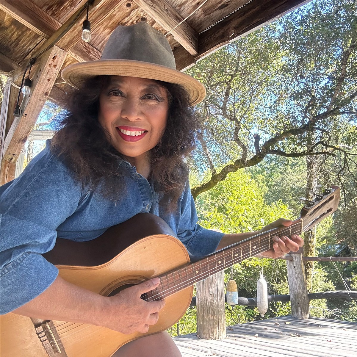 Michelle smiling with her acoustic guitar on a sunlit porch surrounded by trees